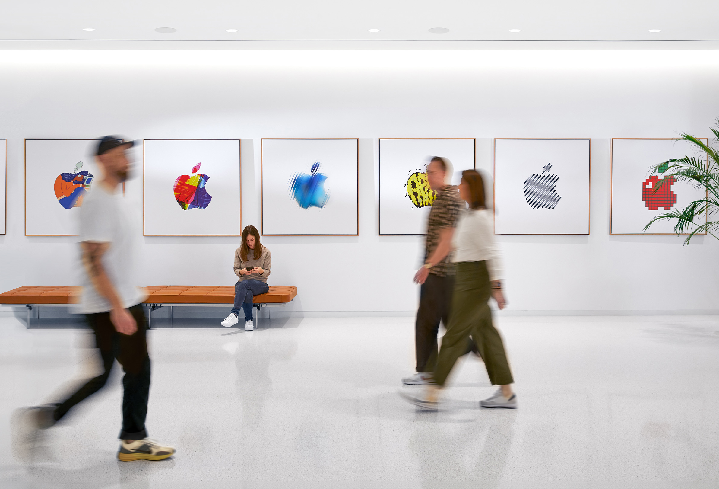 Üç Apple çalışanı renkli Apple logolarının yer aldığı bir iç mekan duvarının önünden geçerken dördüncü bir Apple çalışanı bankta oturuyor.