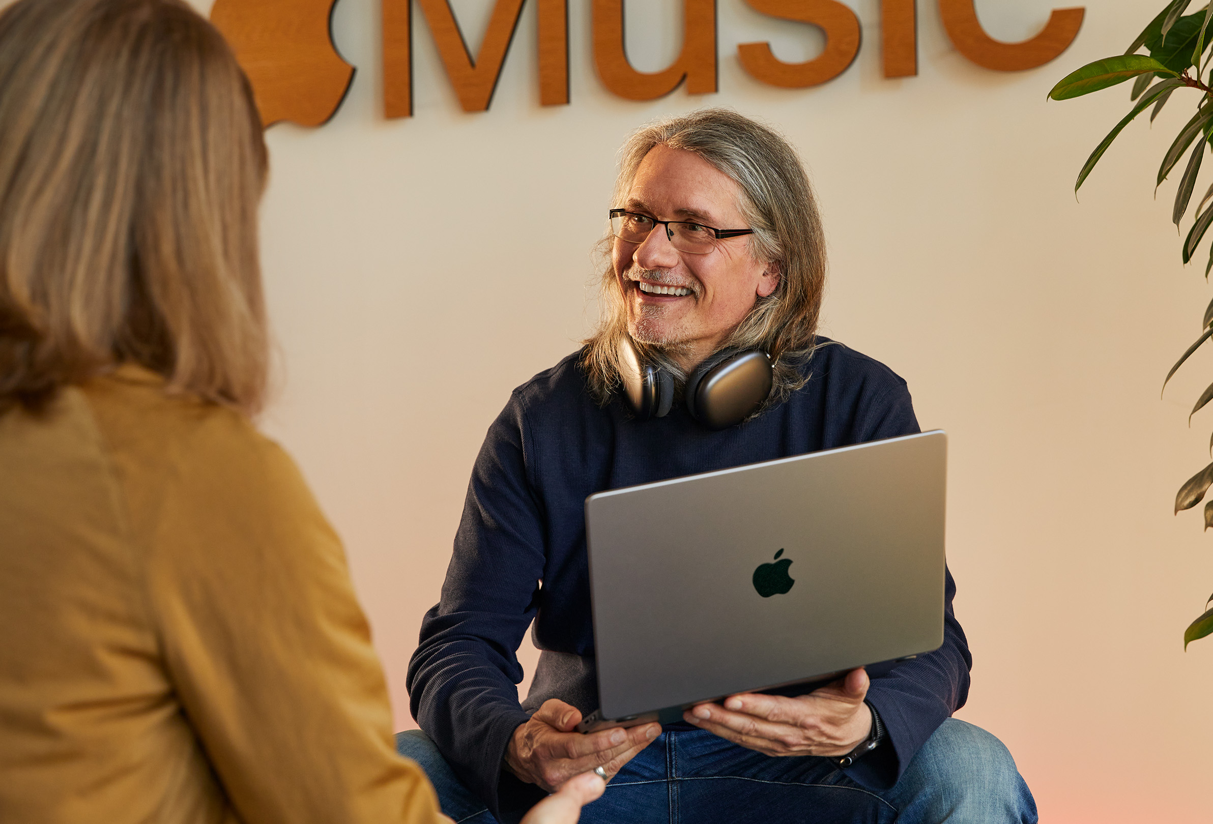 Two Apple employees in conversation, one holding a MacBook and sitting in front of a wall with the Apple Music logo on it.