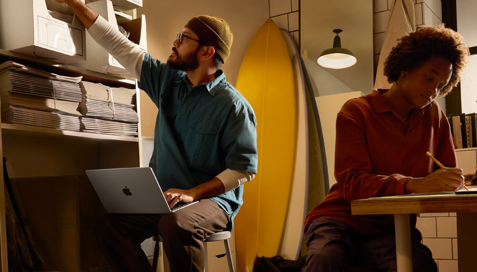 Person in einer Arbeitsumgebung, die auf einem Stuhl sitzt und ein MacBook Air auf ihrem Schoß nutzt, das nicht am Strom angeschlossen ist. Mit der anderen Hand greift sie nach einer Schachtel auf einem hohen Regal.